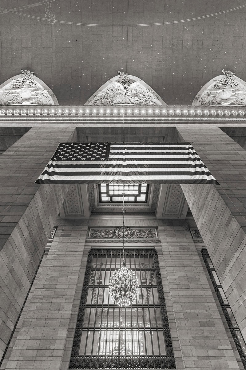 An American flag hangs proudly between high-rise shadows in New York City—an iconic symbol caught in a quiet, windless moment by Dan Kosmayer.