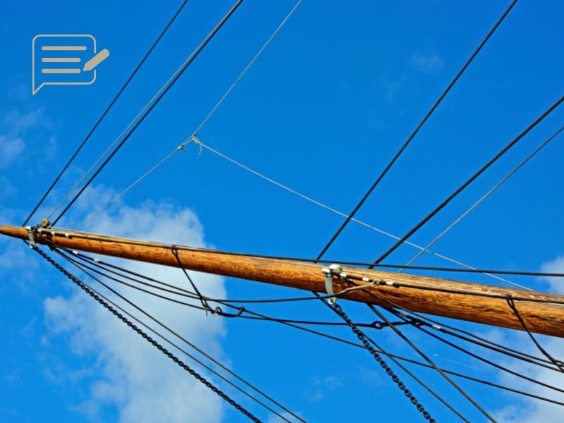 The bowsprit of the Cutty Sark, extending forward, supported by a network of ropes and rigging against a bright blue sky. A classic element of this historic London landmark.
